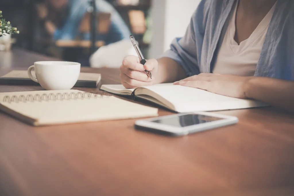 Person writing a will in a notebook with a smartphone and coffee on the desk
