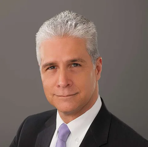 Man with silver hair in a suit and lavender tie, against a gray background.