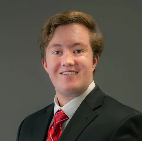 Ryan Welsh in a suit with a red tie, smiling, against a plain gray background.