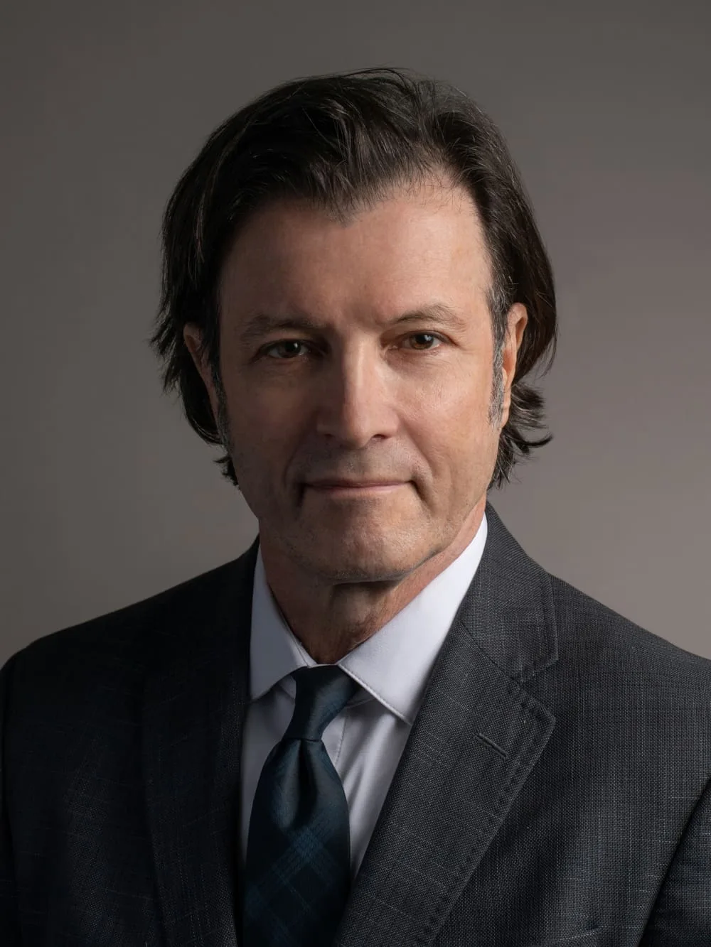Man in a suit and tie with dark hair, against a neutral background.