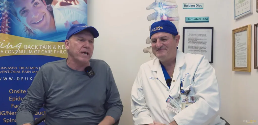 Two men, one in medical attire, sit and talk in a clinical setting with spine-related posters.