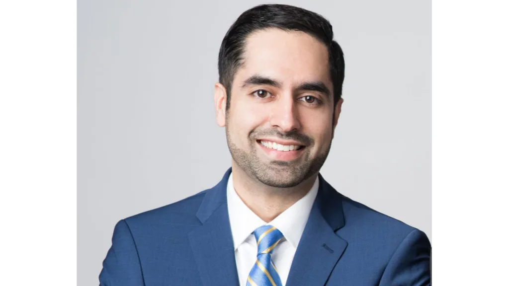 Smiling man in a blue suit with a striped tie against a gray background.
