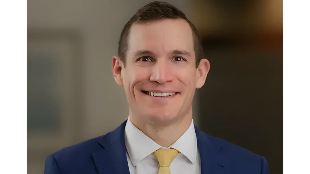Smiling man in a blue suit and yellow tie against a blurred background.