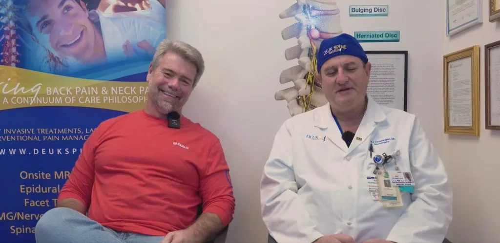 Two men sitting, one in a red shirt and the other in a white lab coat, with medical posters in the background.