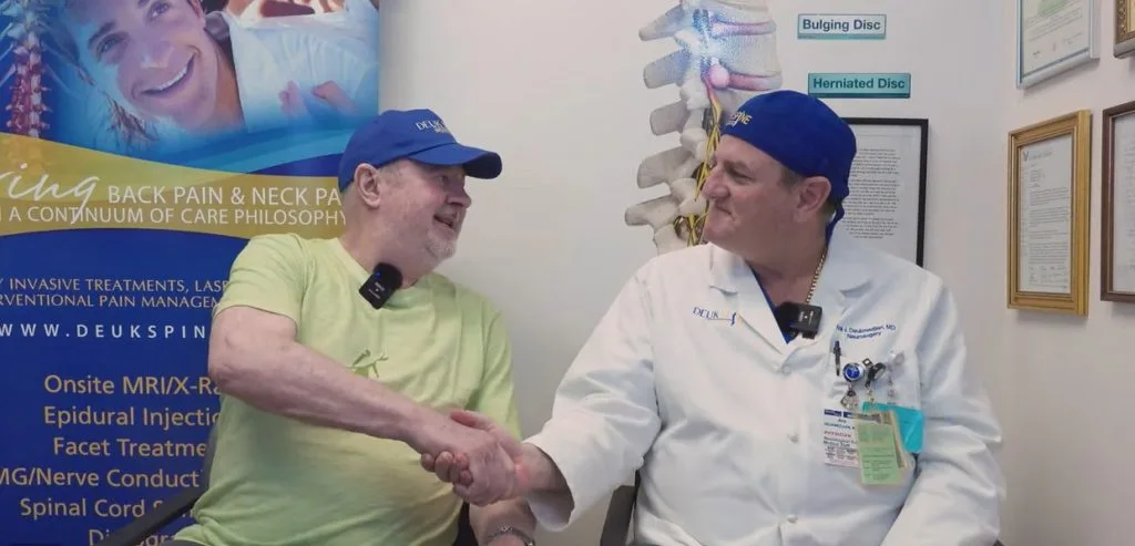 Two men in medical attire shake hands, smiling, with informational posters in the background.
