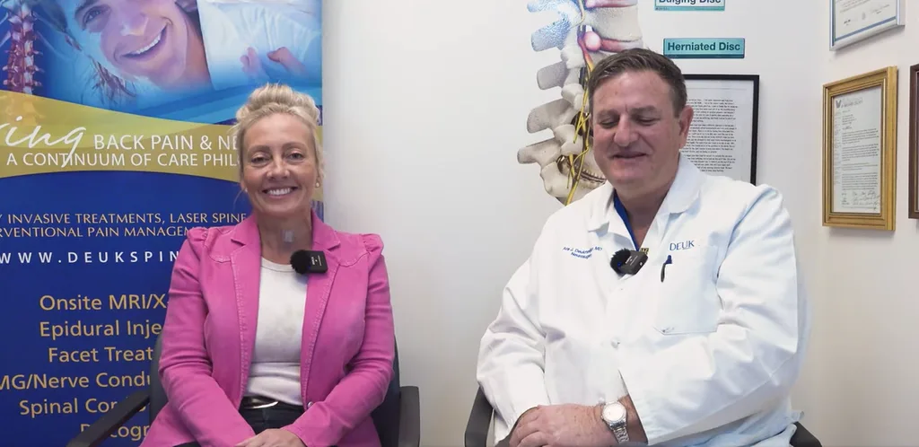 A woman in a pink blazer and a man in a white coat sit smiling in an office with spine posters.