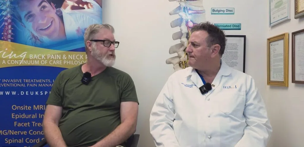 Two men seated in discussion, with medical certificates and spine diagrams on the wall behind them.