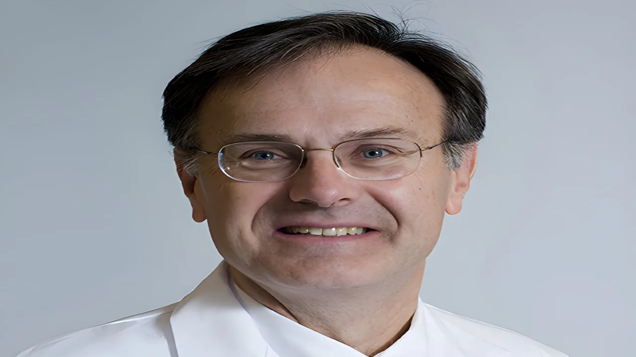 Smiling man wearing glasses and a white coat, against a plain background.