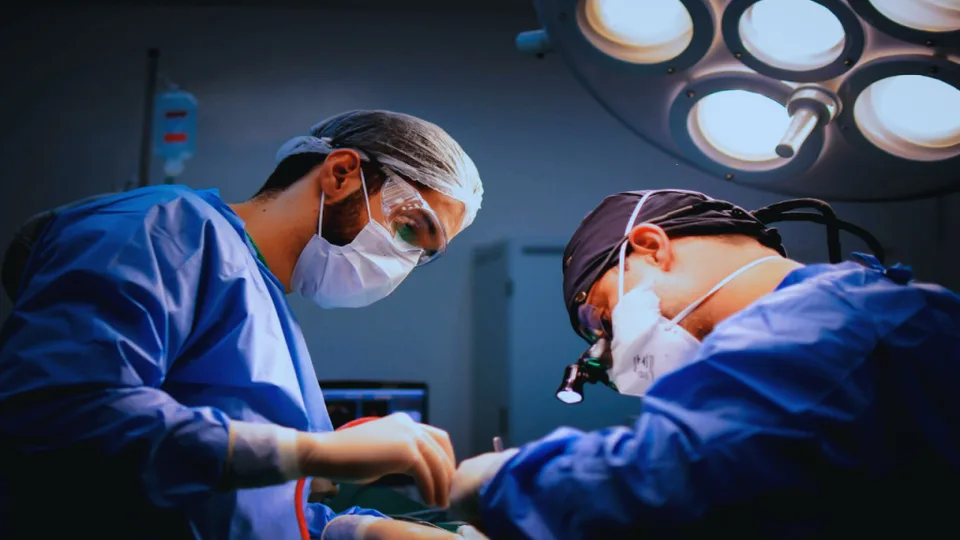 Two surgeons in masks and scrubs perform surgery under operating room lights.