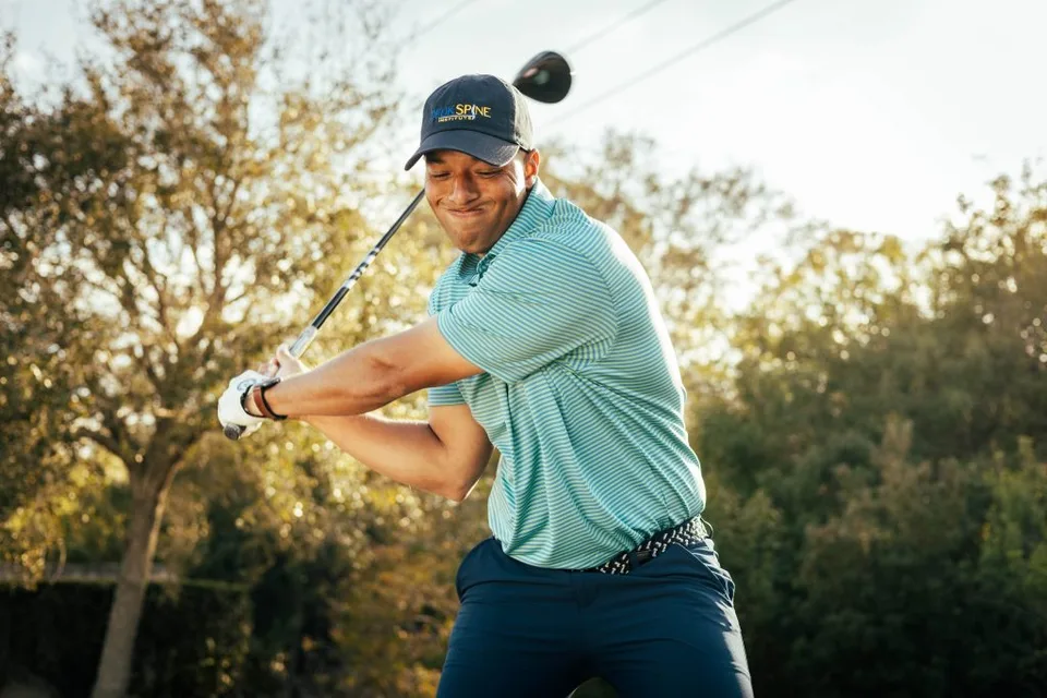 Golfer in mid-swing with a hat, surrounded by sunlight and trees in the background.