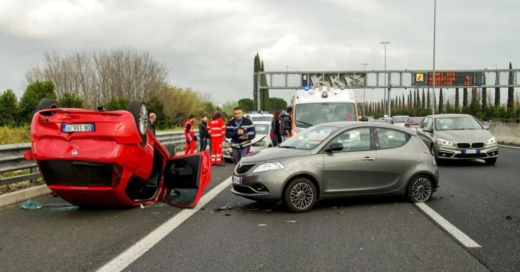 A red car flipped upside down on a highway after a serious collision with emergency responders and an ambulance at the scene