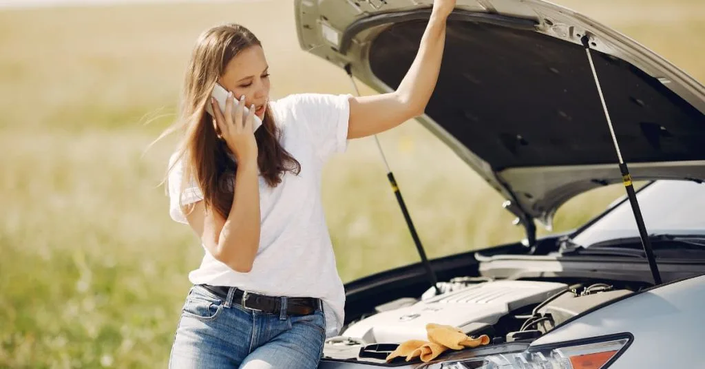 Woman standing by her broken-down car with the hood open, calling for emergency roadside assistance on her phone after experiencing vehicle trouble on the road