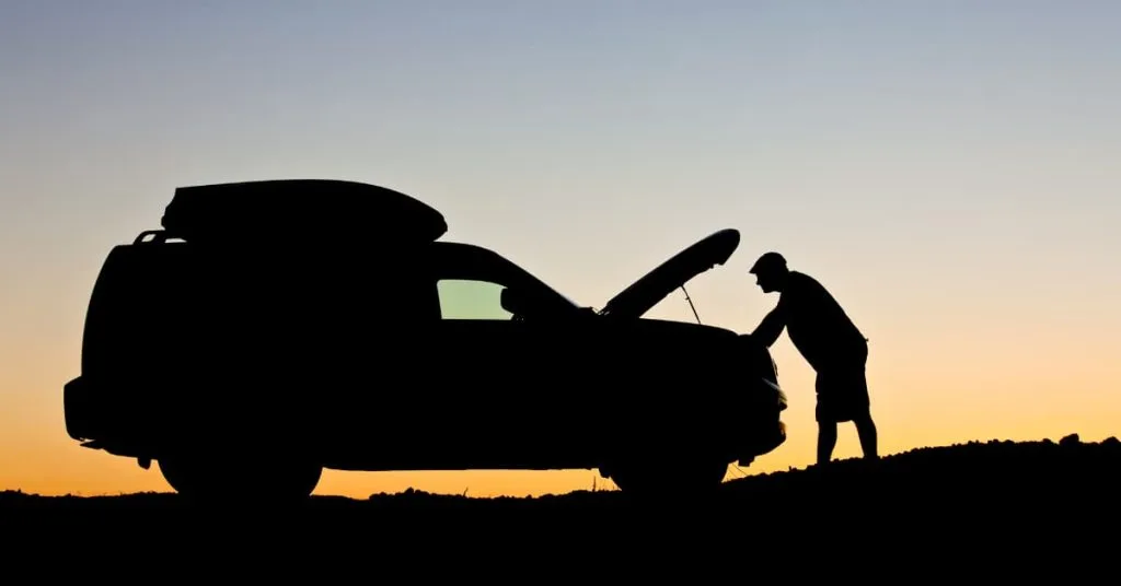 24/7 Roadside Assistance in Maryland: Get Help Now 2 Silhouette of a person checking under the hood of a car at sunset, waiting for 24/7 roadside assistance during a breakdown.