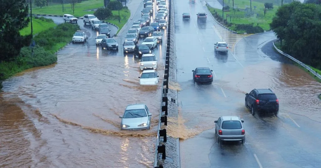 Vehicles drive cautiously through deep floodwater covering a busy roadway during heavy rain, highlighting dangerous conditions that may require towing or roadside assistance.