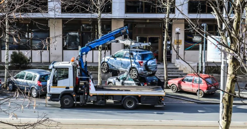 Can You Legally Stop Your Car From Being Towed? 1 Tow truck with a mounted crane lifting a blue SUV onto its flatbed on a busy city street, surrounded by parked cars and buildings, symbolizing vehicle towing or impound.