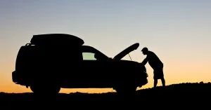 Silhouette of a person checking under the hood of a car at sunset, waiting for 24/7 roadside assistance during a breakdown.