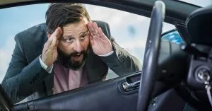 Stressed man in a suit looking through a car window with hands on his head, realizing he is locked out of his vehicle with the keys inside.