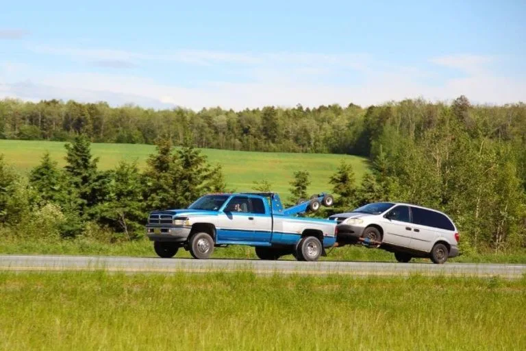 Blue and silver tow truck using a tow dolly to transport a disabled minivan along a scenic countryside highway with green fields and trees in the background