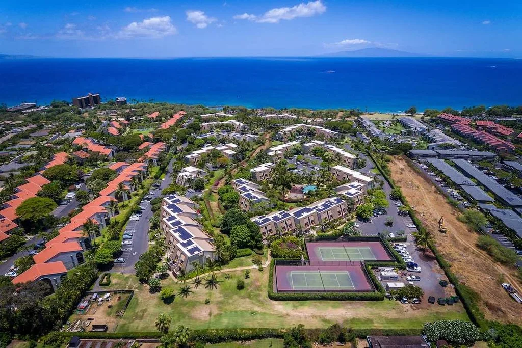 Maui Kamaole Condos for Sale 1 An aerial picture looking over the Maui Kamaole Condos towards the bright blue Pacific Ocean. It is a sunny day with a few white puffy clouds. The Island of Lanai can be seen in the distance. This perspective of Maui Kamaole shows the tennis courts on the property and you can see one of the swimming pools. Solar panels can be seen on the roofs of many of the buildings.