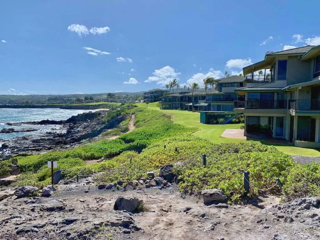 This photo shows condominiums along the front row of Kapalua Bay Villas. A rocky shoreline is in the foreground and you can see Oneloa Beach in the distance
