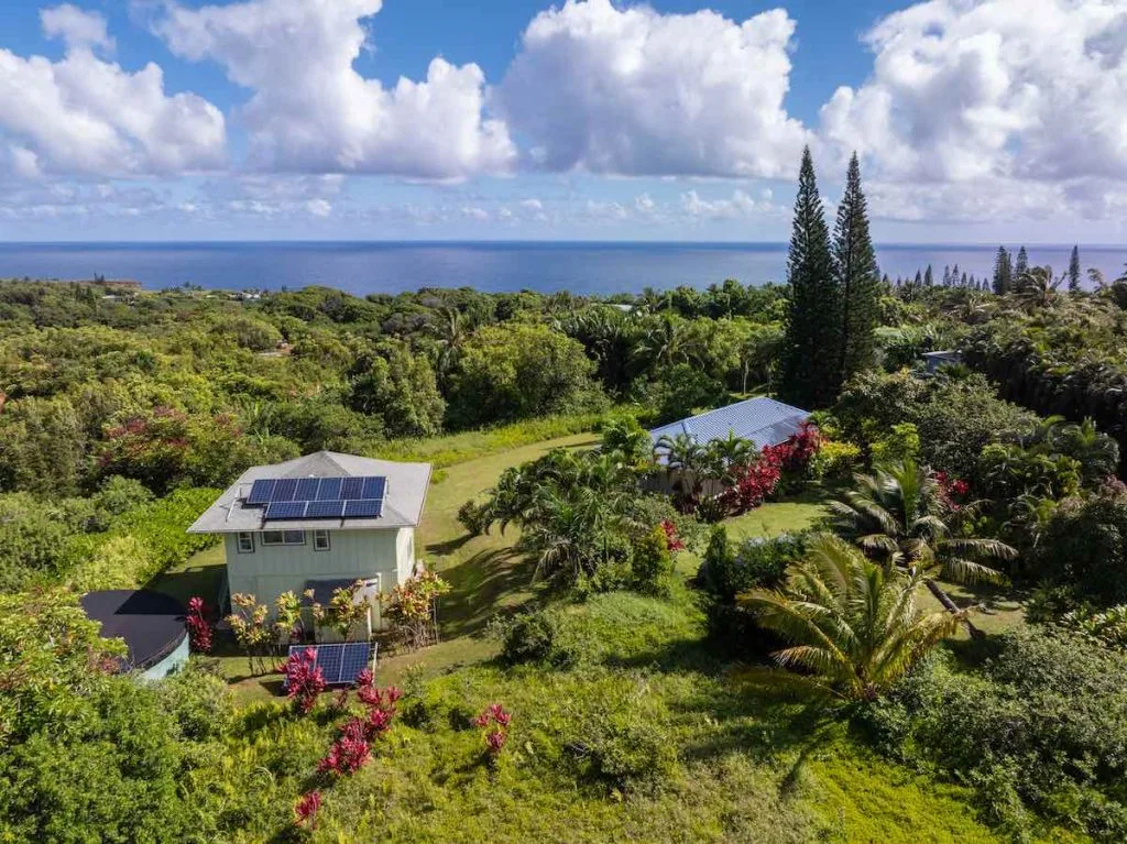 This photo shows an aerial image of an off the grid home with a detached garage and ag office set in a lush landscape of palm trees, cook island palms, vibrant red ti plants. A  vibrant blue ocean is close by. Fluffy white clouds carried by the trade winds bring occasional showers. 