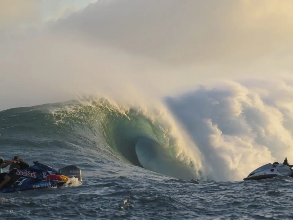 A massive ocean wave forming a hollow tube with two jet skis visible on the surface, one carrying a Red Bull logo, under a sky with soft lighting.