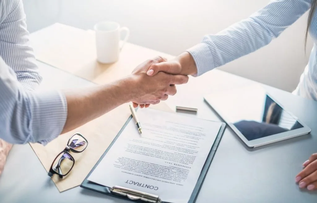 Two people shaking hands over a signed contract on a desk with glasses, pen, and tablet, symbolizing agreement and partnership in legal services.
