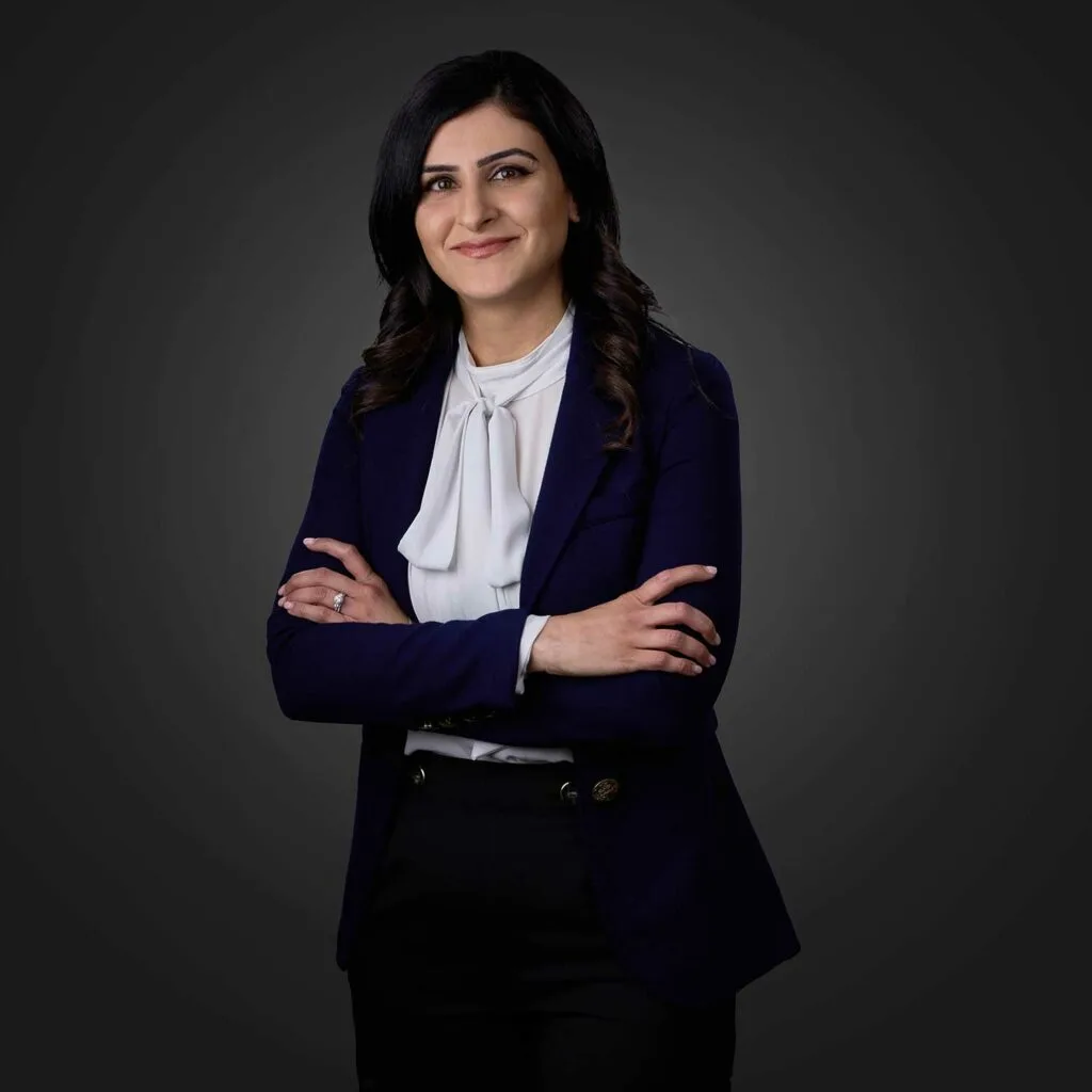 Smiling woman with long dark hair wearing a white blouse with a bow and a dark navy blazer, standing with arms crossed against a dark grey background.