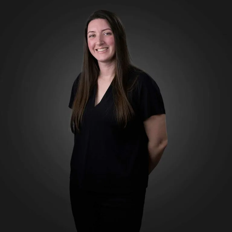 Smiling woman with long straight brown hair wearing a black short-sleeve V-neck top and black pants standing against a dark gray background with her hands behind her back