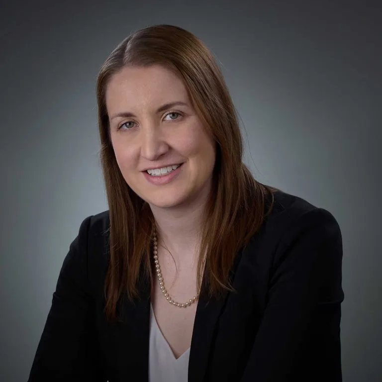 Smiling woman with straight brown hair wearing a black blazer, light blouse, and a pearl necklace against a gray gradient background