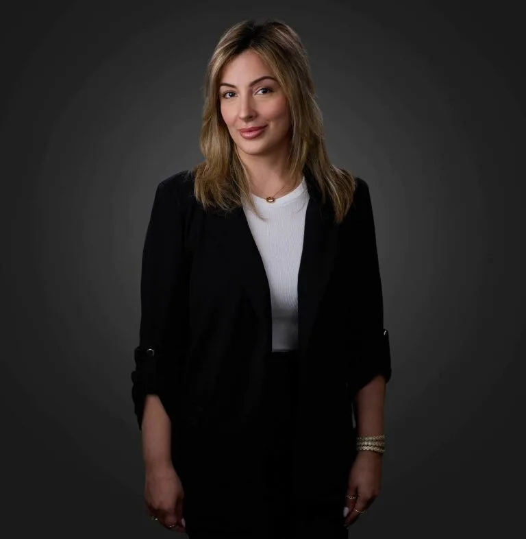 Smiling woman with light brown hair wearing a black blazer and white top, standing against a dark gray background