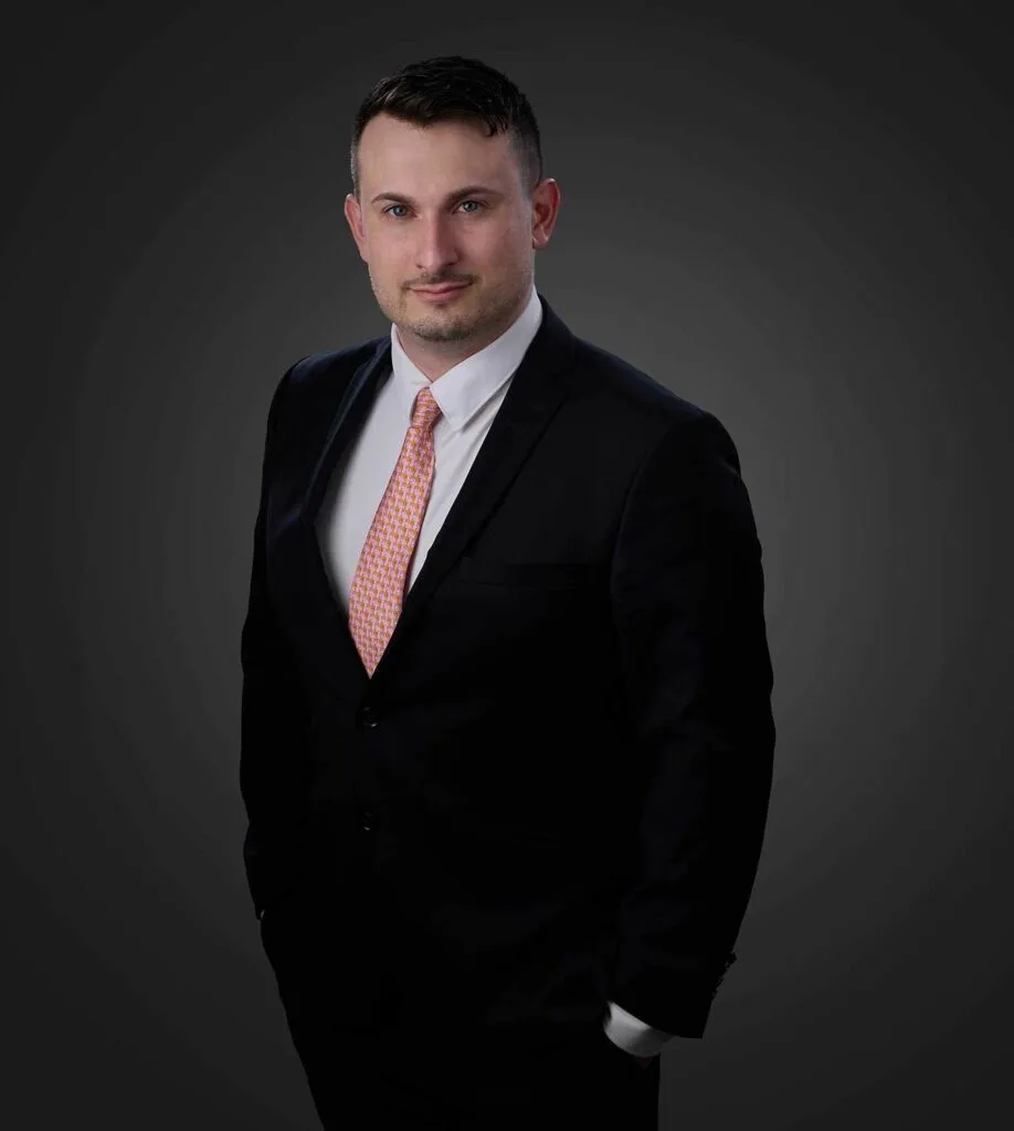 Man in a black suit, white dress shirt, and patterned orange tie standing against a dark gray background, looking at the camera with hands in pockets.