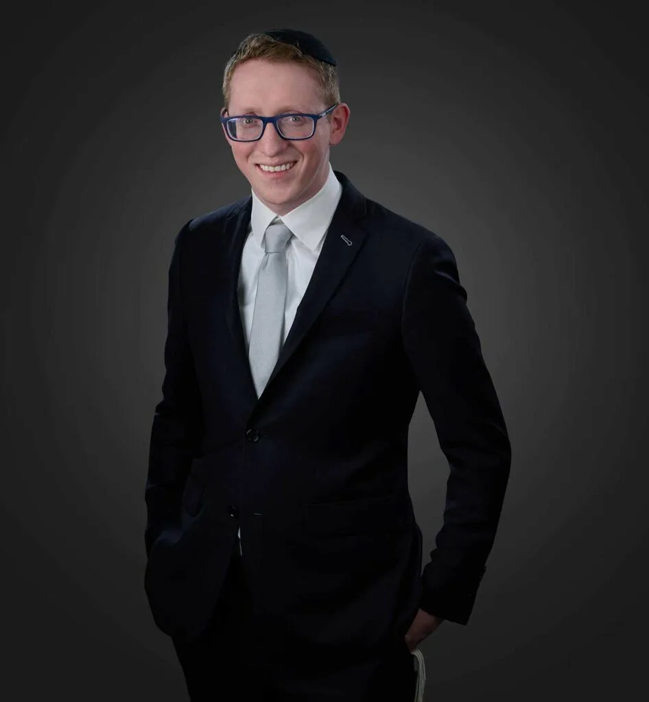 Smiling man wearing a dark suit with a light gray tie, blue glasses, and a black kippah, standing against a dark gray background with hands in pockets.