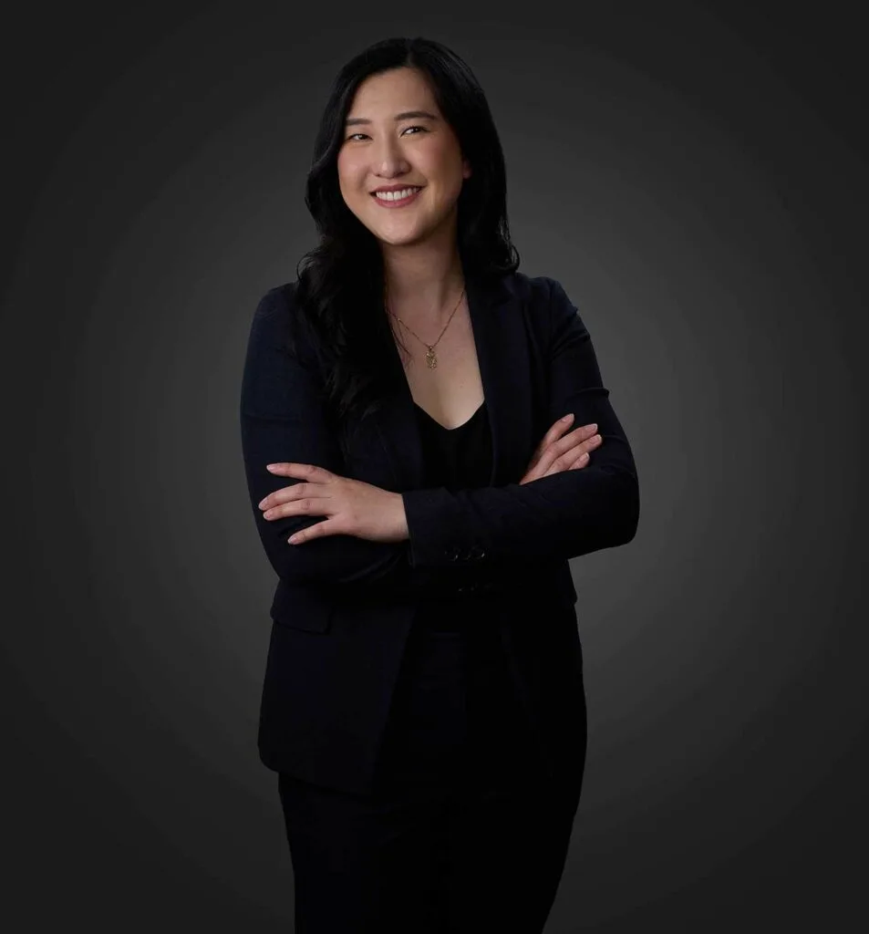 Smiling woman with long black hair wearing a black blazer and top, standing with arms crossed against a dark gradient background.