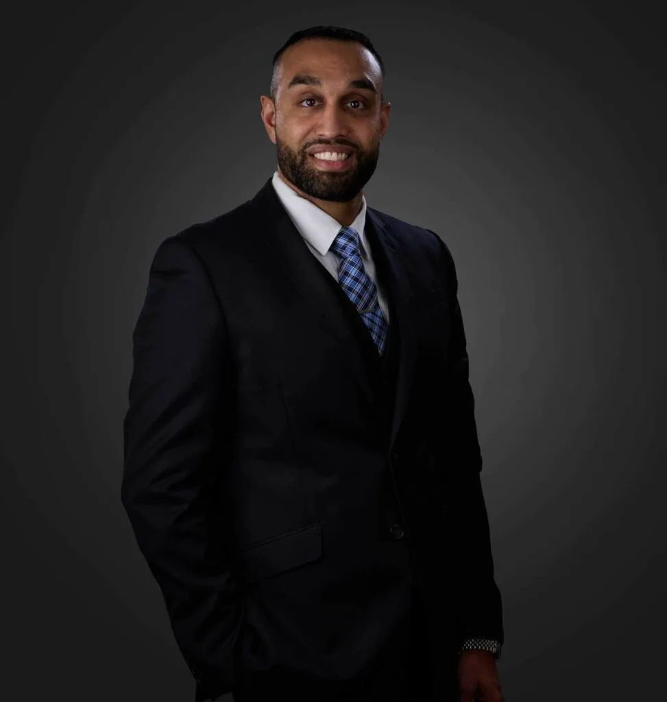 Smiling man with a beard wearing a black suit, white shirt, and blue checkered tie, standing against a dark gray gradient background with one hand in his pocket.