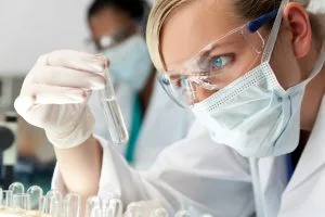 Blonde woman in a lab setting looking at a clear tube while wearing protective glasses, gloves and a face mask