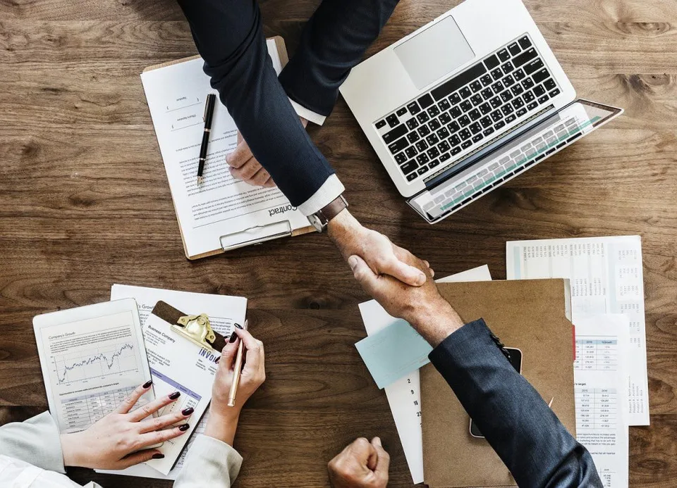 two people shake hands over desk and laptop