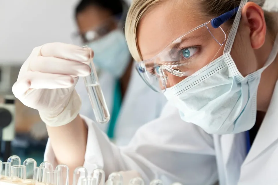 Blonde woman in a lab setting looking at a clear tube while wearing protective glasses, gloves and a face mask