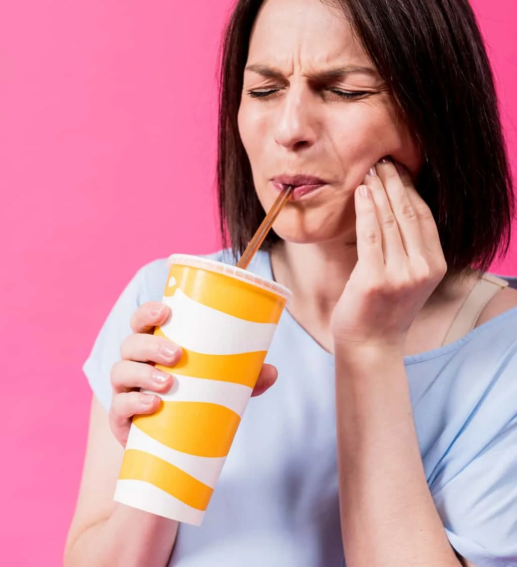 gum recession signs: recognizing the early warnings your mouth is sending 2 woman drinking from a cup while holding her cheek in discomfort against a pink background.