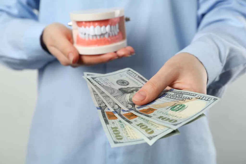 dental implants vs dentures cost, comfort, and long-term value 2 hands holding a denture model beside a stack of us hundred-dollar bills