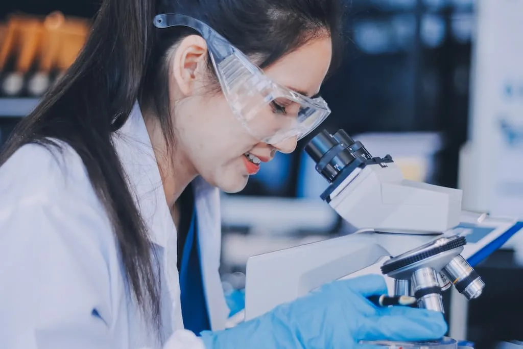 scientist wearing safety goggles examines a sample under a microscope in a lab.