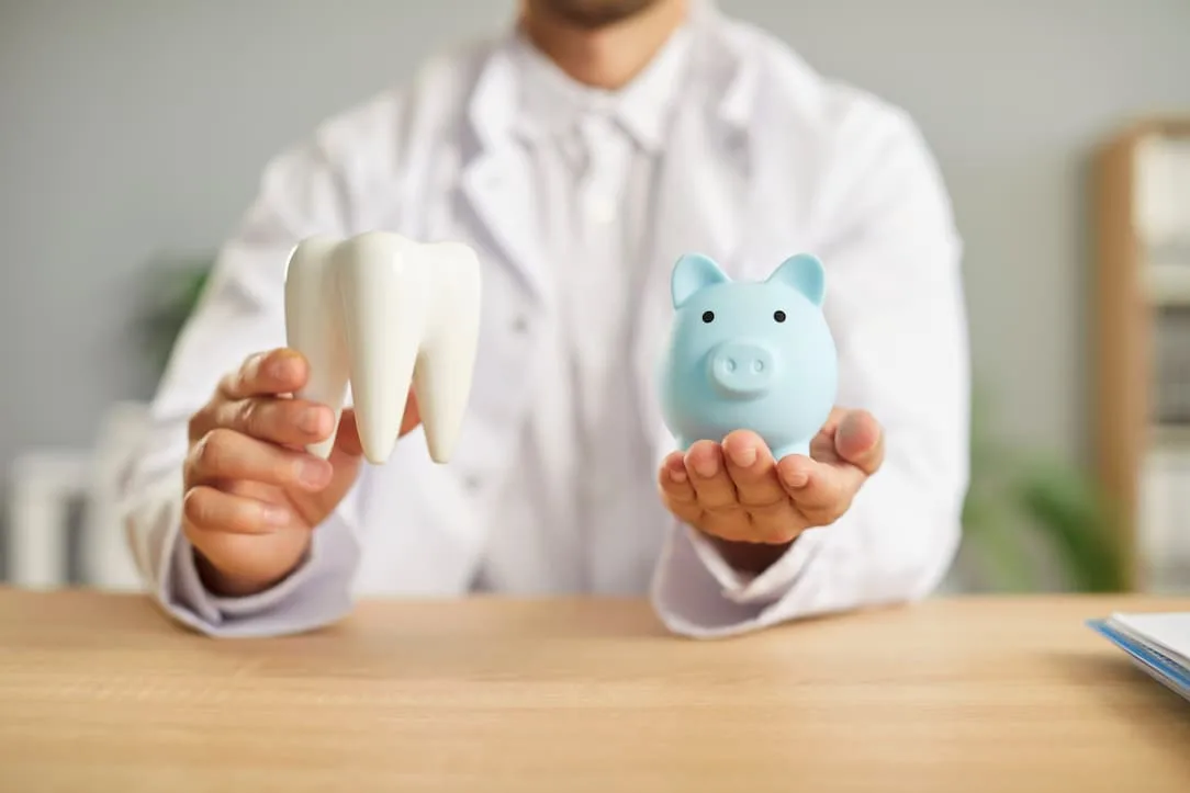 person in a white coat holding a large tooth model and a blue piggy bank.