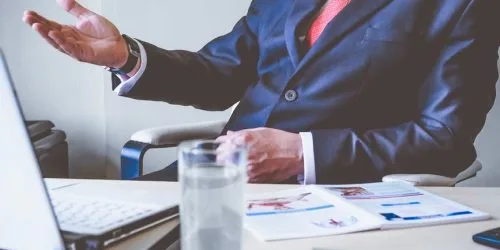 Business professional in a suit discussing ideas at a desk with a laptop and documents. Water glass in the foreground.