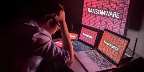 A person stressed by ransomware alerts on multiple computer screens in a dark setting, illustrating cyber security threats.
