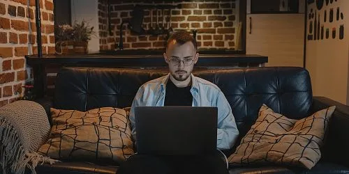 A young man working on his laptop, sitting on a cozy couch in a stylish, brick-walled living space.