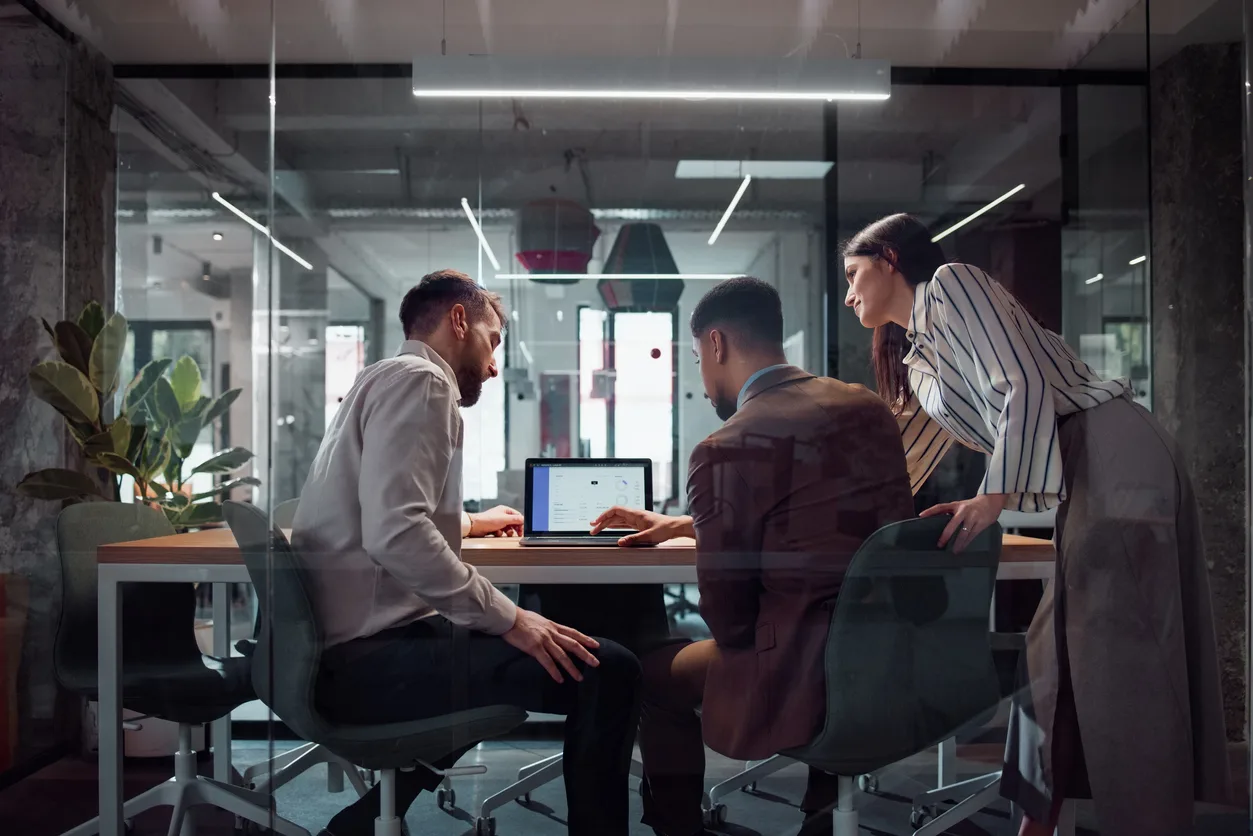Three colleagues collaborating in a modern office, reviewing data on a laptop at a meeting table.
