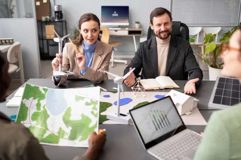 Group of professionals discussing renewable energy project using wind turbine models, solar panel samples, and environmental maps in a modern office setting