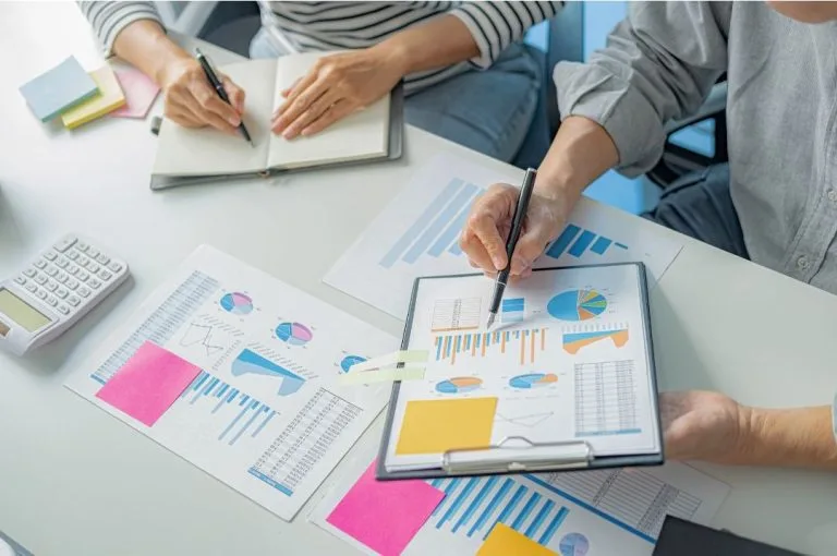 Two individuals examining financial graphs and data charts on papers spread out on a desk during a strategy meeting.