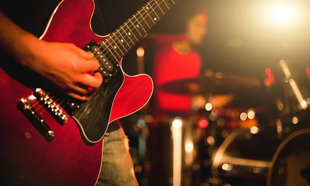 A red guitar played by someone in a high school rock band in blue jeans and a black T-shirt, with a drummer and drum set in the background.