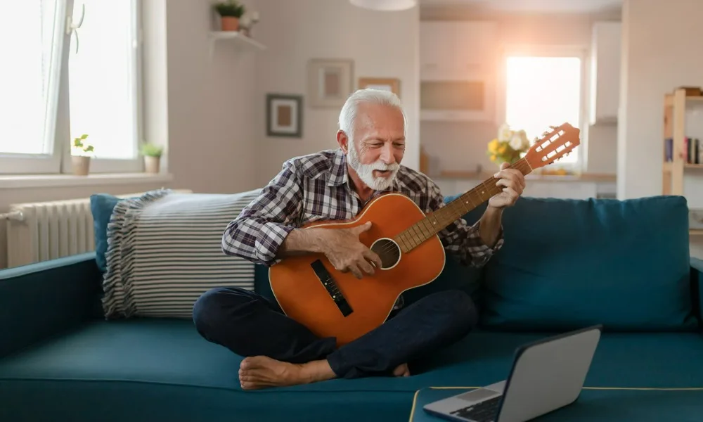 The Trick to Learning Guitar as an Adult 15 Retired man with gray hair sitting barefoot on a couch, learning to play guitar as an adult virtually with a computer in front of him.